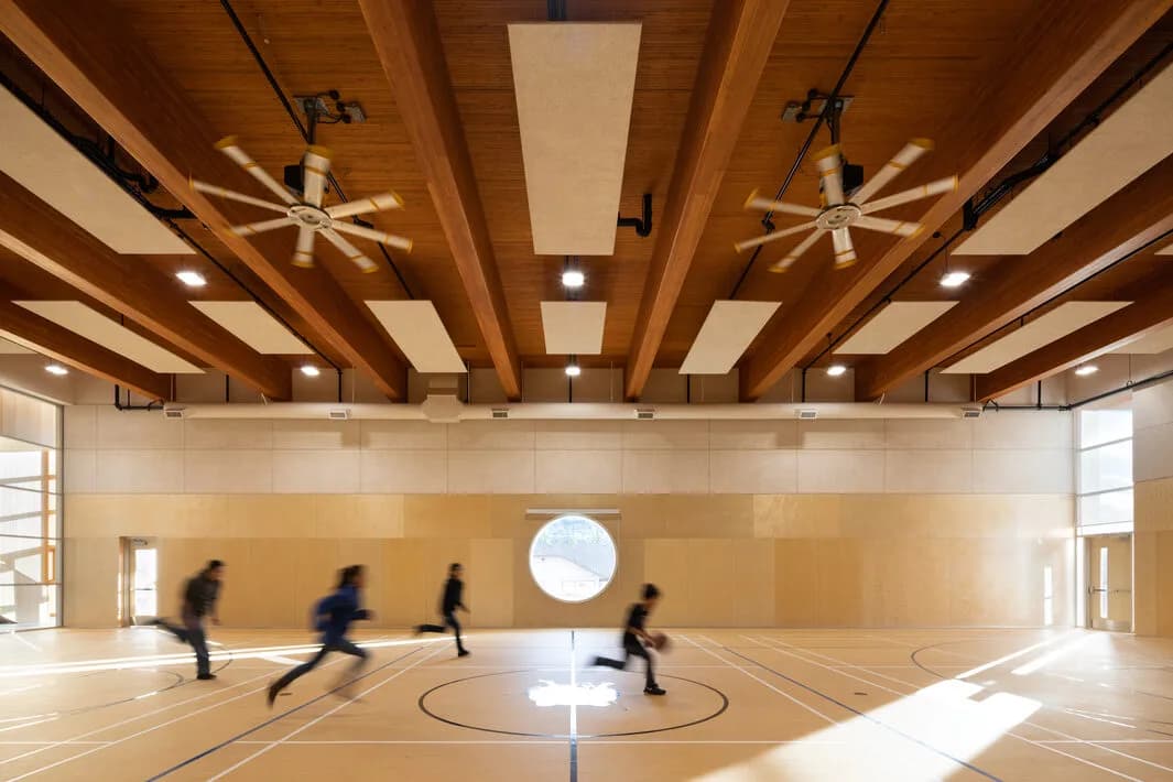 Children playing basketball in the gymnasium beneath exposed glulam beams and NLT panels, with a round window looking out.